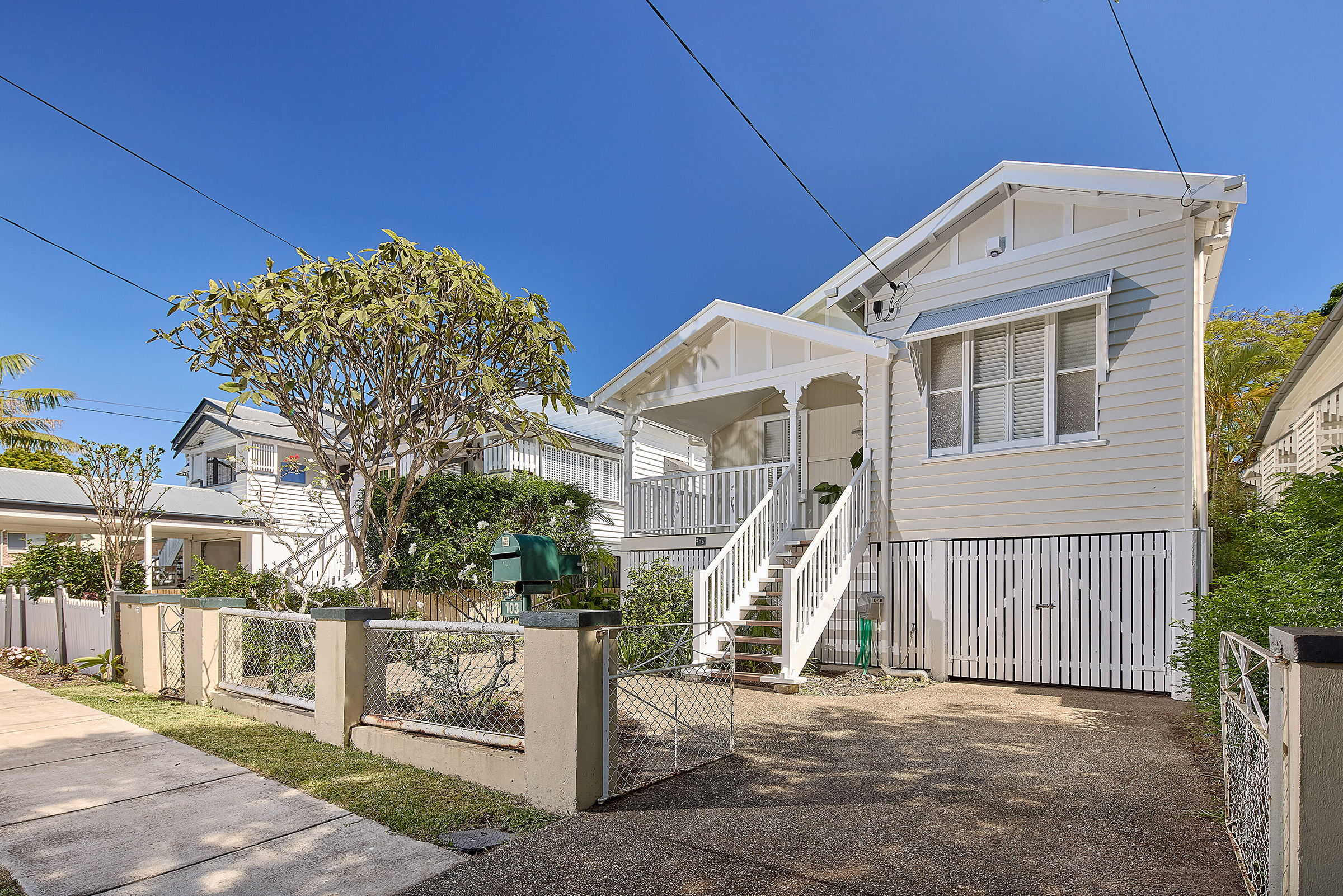 Classic white elevated home with front stairs, veranda, and driveway behind a front fence.