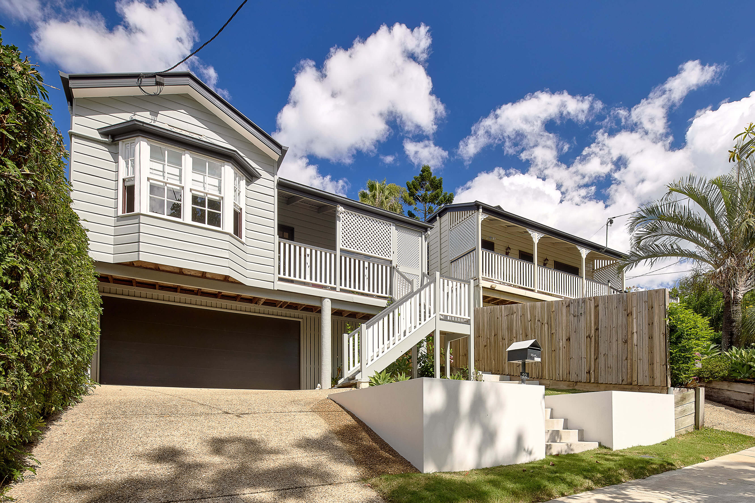Elevated grey home with front stairs, wraparound veranda, and double garage under a bright blue sky.
